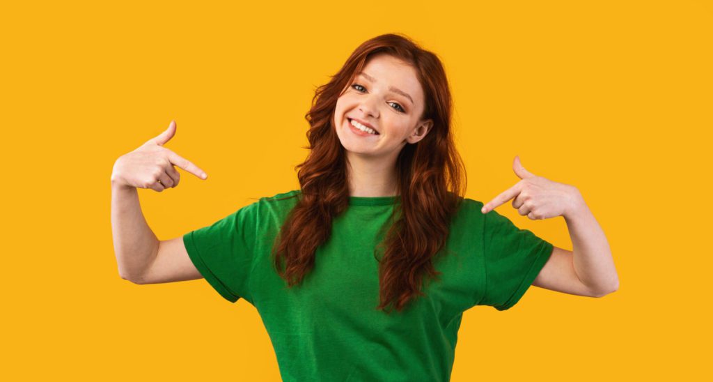 Smiling Red Haired Girl Pointing Fingers At Herself Over Yellow Background